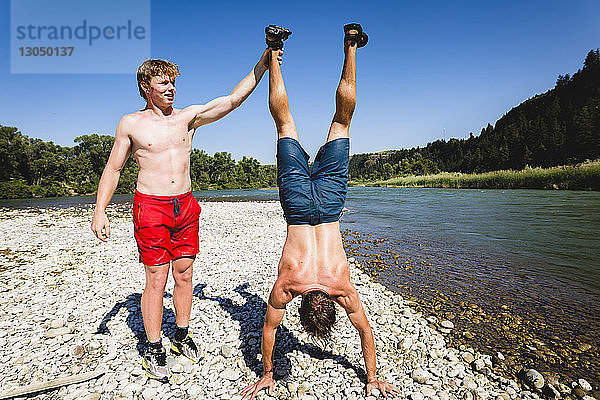 Teenager hilft Freund beim Handstand am Seeufer im Sommer