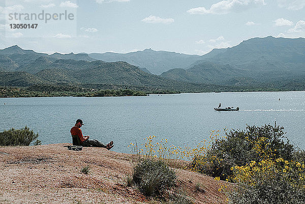 Seitenansicht eines Mannes  der am Seeufer vor Bergen sitzt