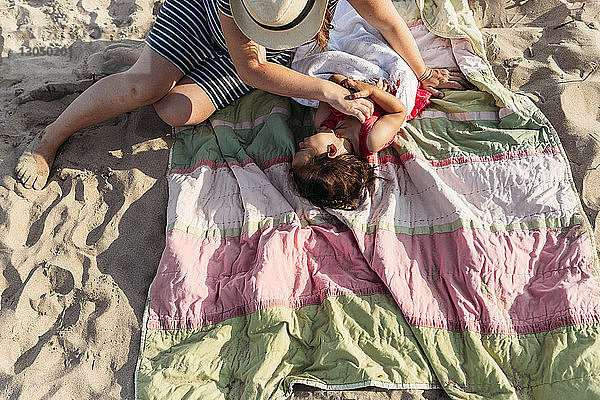 Hochwinkelaufnahme einer Mutter  die ihre Tochter kitzelt  die auf einer Picknickdecke am Strand liegt