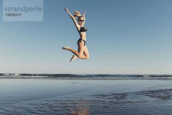 Unbeschwerte Frau springt am Strand gegen klaren Himmel