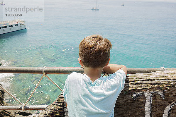 Rückansicht eines Jungen  der am sonnigen Tag an der Reling aufs Meer schaut