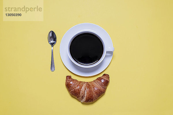 Draufsicht auf schwarzen Kaffee  Löffel und Croissant auf gelbem Tisch