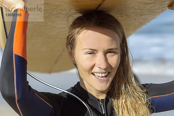 Porträt einer fröhlichen Frau mit Surfbrett auf dem Kopf am Strand