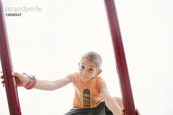 Niedrigwinkel-Porträt eines Jungen auf einem Spielgerät im Freien vor klarem Himmel