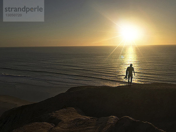 Scherenschnitt eines Mannes mit Surfbrett  der bei Sonnenuntergang auf einer Klippe am Meer steht