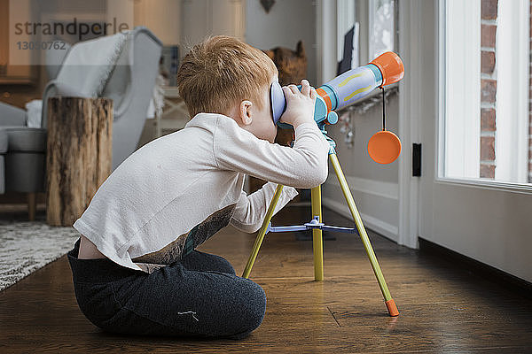 Seitenansicht eines Jungen  der durch ein Teleskop schaut  während er zu Hause an der Tür sitzt