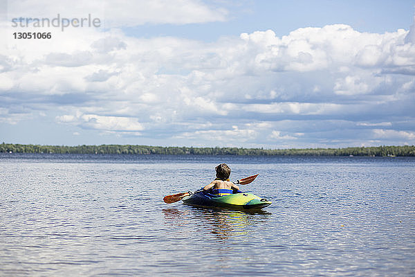 Rückansicht eines Kajak fahrenden Jungen auf einem See vor bewölktem Himmel