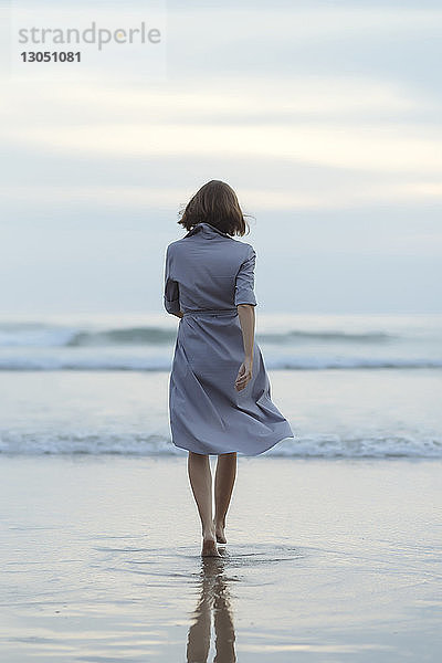Rückansicht einer sorglosen Frau  die bei Sonnenuntergang am Strand in Richtung Meer geht