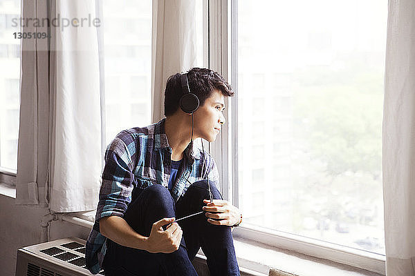 Nachdenklicher Mann hört Musik  während er zu Hause durchs Fenster schaut