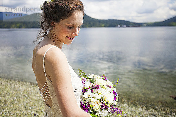 Seitenansicht der am Seeufer stehenden lächelnden Braut mit Blumenstrauss