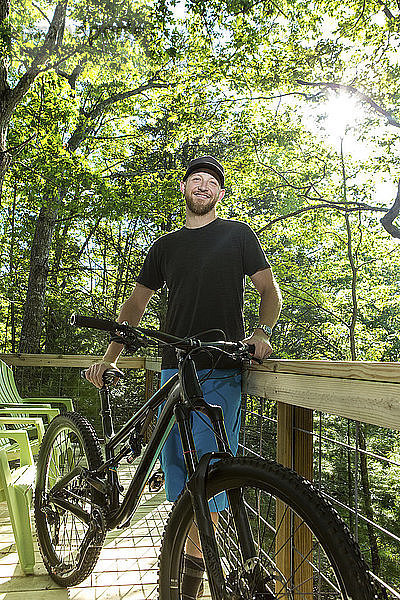 Porträt eines selbstbewussten Wanderers  der ein Mountainbike in der Hand hält  während er am Zaun eines Campingplatzes im Wald steht