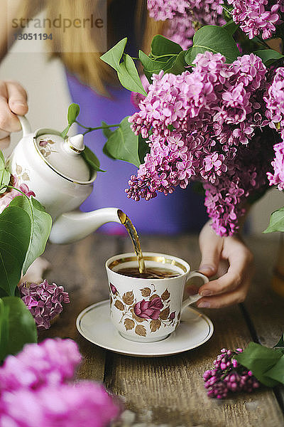 Mitschnitt einer Frau  die auf einem Holztisch Tee in einer Tasse neben Blumen ausschenkt