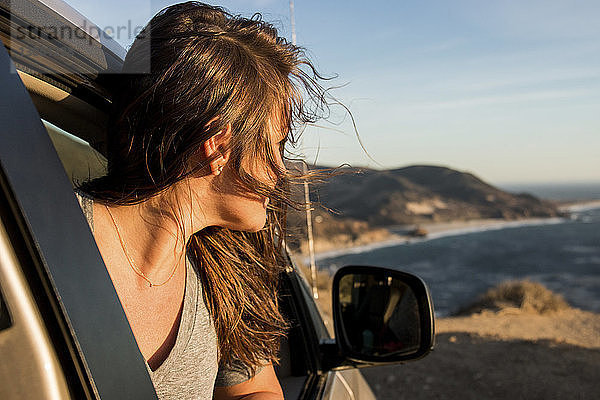Frau  die bei Sonnenuntergang durch ein Autofenster am Strand gegen den Himmel schaut
