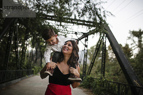 Fröhliche Mutter trägt Sohn auf den Schultern an der Brücke