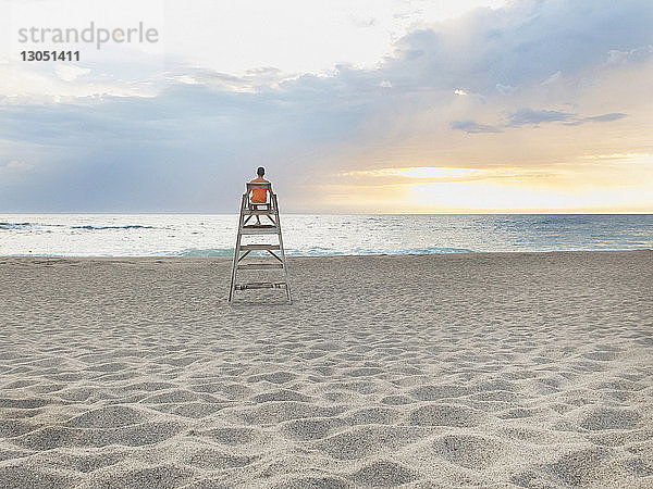 Rückansicht eines Mannes  der auf einer Bademeisterhütte am Strand vor bewölktem Himmel bei Sonnenuntergang sitzt