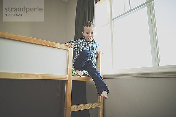 Ganzflächiges Porträt eines lächelnden Jungen  der zu Hause auf einem Etagenbett am Fenster sitzt