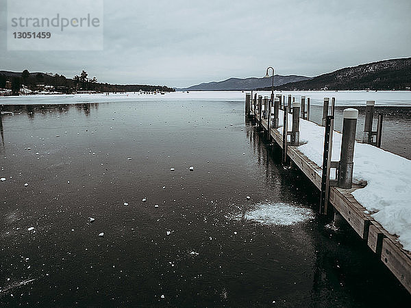 Schneebedeckter Pier über gefrorenem See vor bewölktem Himmel