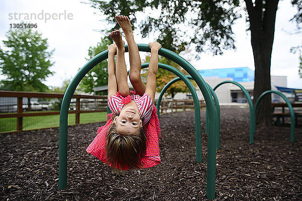 Porträt eines Mädchens  das kopfüber auf einem Spielgerät im Freien auf dem Spielplatz hängt