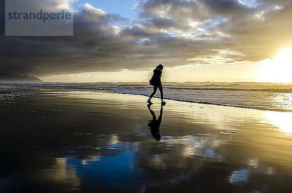 Silhouette einer Frau  die bei Sonnenuntergang am Strand vor bewölktem Himmel spazieren geht