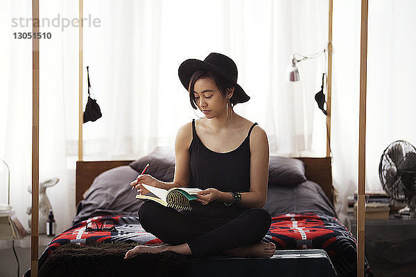 Frau liest zu Hause auf dem Bett sitzend ein Buch
