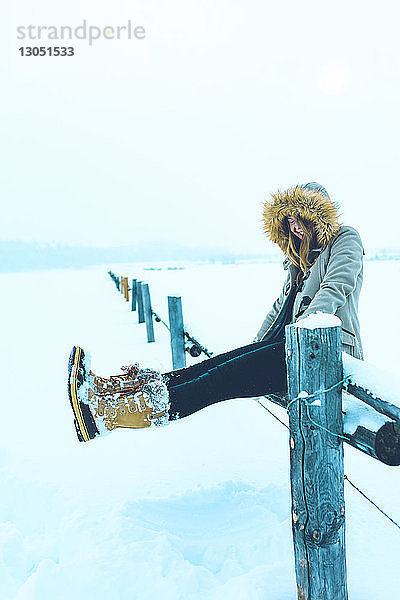Fröhliche Frau sitzt im Winter auf dem Geländer eines schneebedeckten Feldes