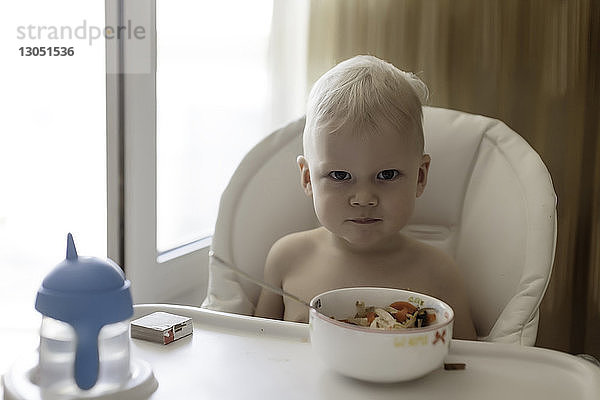 Porträt eines Jungen ohne Hemd  der zu Hause auf einem Hochstuhl sitzt und zu essen bekommt