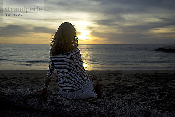 Rückansicht einer Frau  die sich bei Sonnenuntergang am Meer vor bewölktem Himmel entspannt