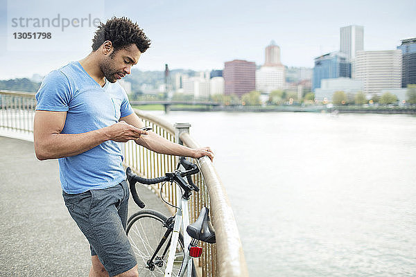 Sportler  der ein Smartphone benutzt  während er mit dem Fahrrad auf einer Brücke am Fluss steht