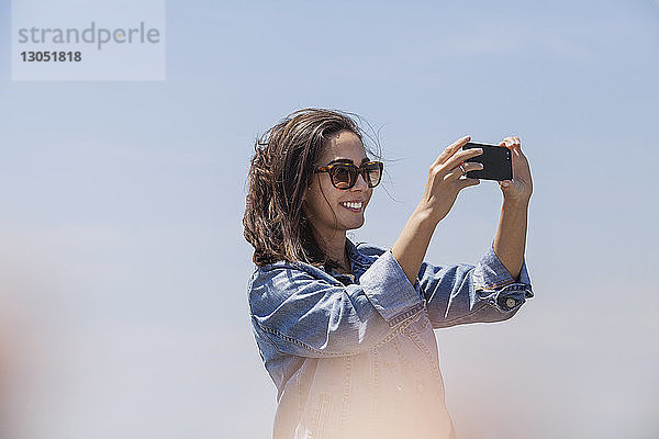 Niedrigwinkelansicht einer glücklichen Frau  die durch ein Smartphone vor klarem Himmel fotografiert