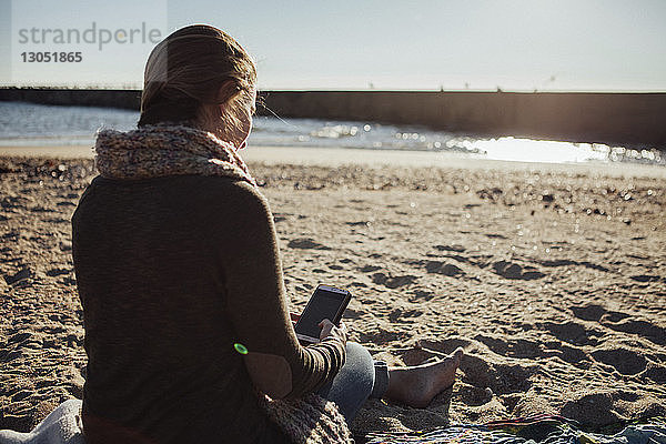 Rückansicht einer Frau  die ein Smartphone benutzt  während sie am Strand im Sand sitzt