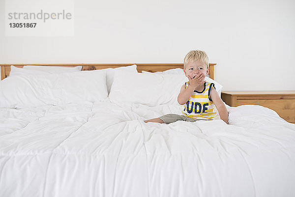 Junge mit der Hand den Mund bedeckend  schaut weg  während er auf dem Bett sitzt