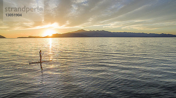 Frau paddelt im Meer gegen bewölkten Himmel bei Sonnenuntergang