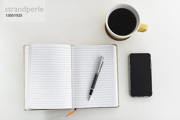Draufsicht von Stift auf Buch bei Kaffeetasse und Smartphone über weißem Hintergrund