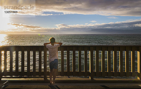 Rückansicht eines Jungen  der auf das Meer schaut  während er bei Sonnenuntergang an der Reling vor bewölktem Himmel steht