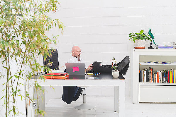 Cooler Geschäftsmann mit den Füßen im Büro beim Blick auf das digitale Tablet