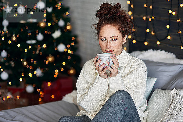 Frau trinkt warmes Getränk auf einem mit Weihnachtslichtern geschmückten Bett