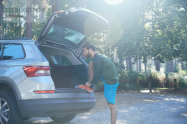 Männlicher Läufer bindet Schnürsenkel am Kofferraum eines Autos im sonnenbeschienenen Wald