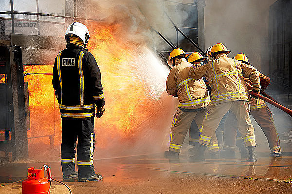 Ausbildung von Feuerwehrleuten  Feuerwehrleute  die Wasser bei einem Brand in einer Trainingsanlage versprühen  Rückansicht