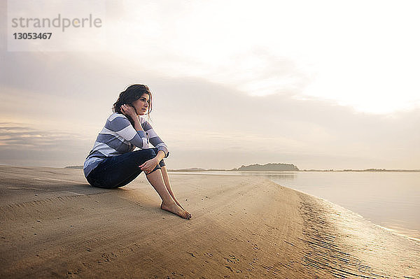 Nachdenkliche Frau sitzt am Meeresufer gegen den Himmel