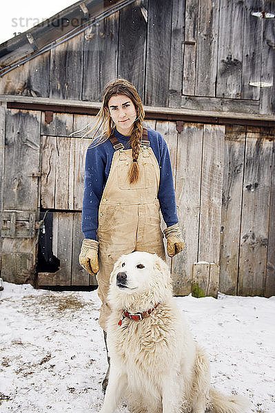 Porträt einer Frau  die neben einem Hund vor einer Scheune auf einem schneebedeckten Feld steht