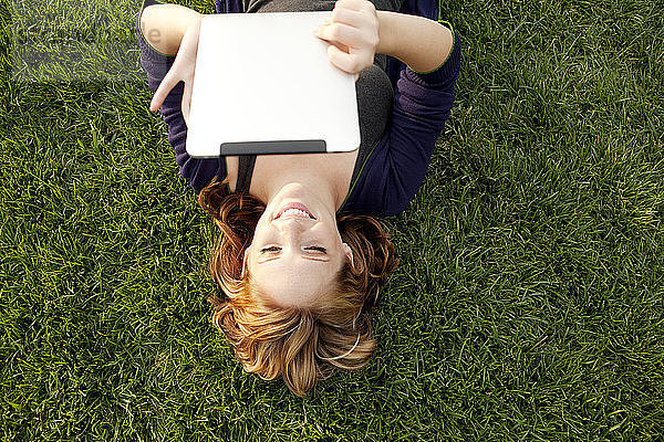 Draufsicht einer glücklichen Frau  die ein digitales Tablett benutzt  während sie auf einem Grasfeld liegt