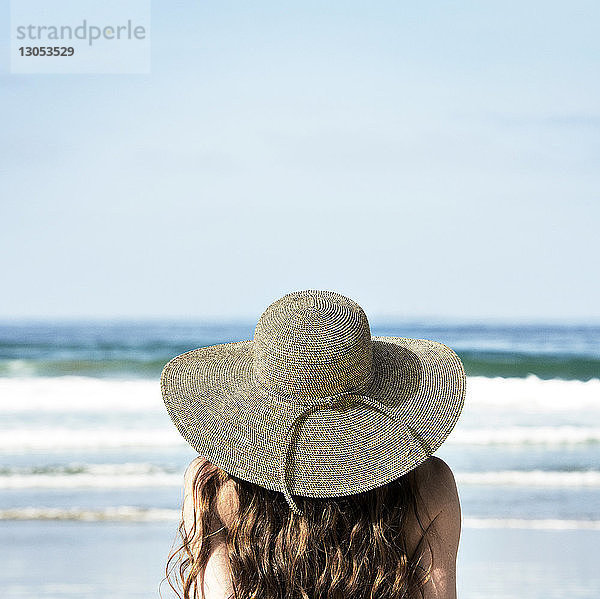 Rückansicht einer am Strand sitzenden Frau mit Sonnenhut