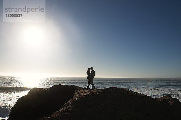 Ehepaar steht auf einem Felsen am Meer gegen den Himmel an einem sonnigen Tag