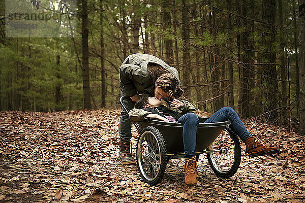 Mann küsst Frau  die im Wald auf einer Schubkarre sitzt
