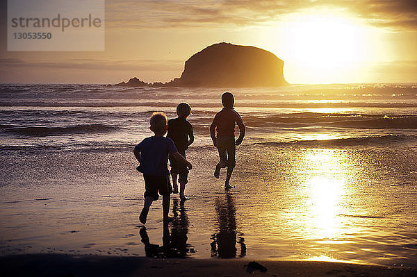 Silhouette von Geschwistern  die bei Sonnenuntergang am Ufer laufen