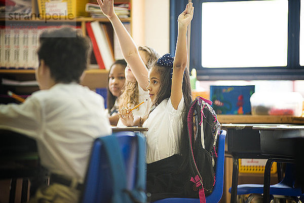 Schulmädchen heben die Hand  während sie im Klassenzimmer sitzen