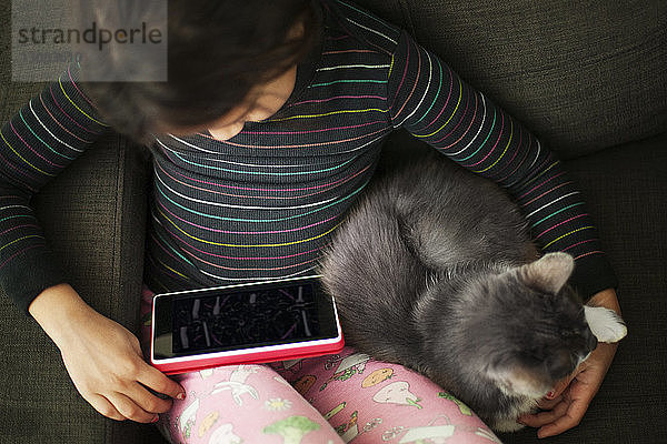 Hochwinkelansicht eines Mädchens  das einen Tablet-Computer benutzt  während es mit einer Katze auf dem Sofa sitzt