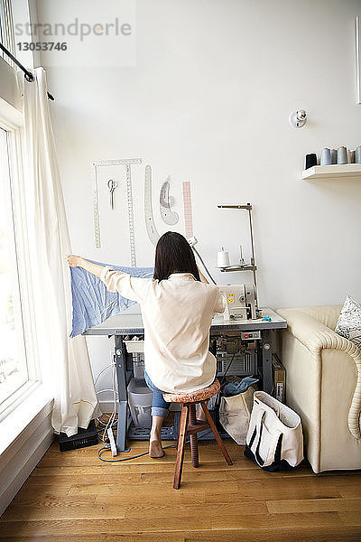 Rückansicht eines Modedesigners  der Stoff hält  während er auf einer Nähmaschine sitzt