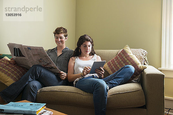 Frau benutzt Tablette von Freundin  die Zeitung liest  während sie zu Hause auf der Couch sitzt