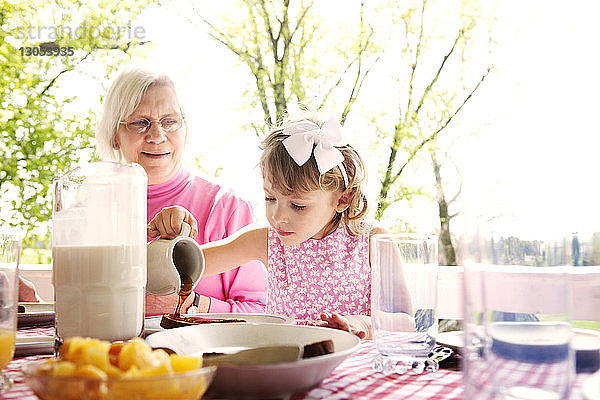 Mädchen gießt Sirup auf das Essen  während die Großmutter am Tisch sitzt